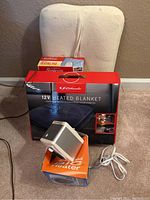Lot photo showing all three items: Schumacher 12V heated blanket in box, Sharper Image heater on orange box, Sunbeam heating pad box behind.