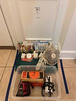 Full lot including three travel irons, clip-on fan, and Glade air fresheners in packaging, shown with carrying plastic bin.