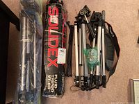 Photo showing all five tripods including Ambico 61 boxed tripod, Solidex Fluid Tripod VT-86 boxed item, and three assorted tripods lying on carpet.