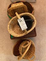 Overview of assorted wicker baskets stacked and grouped, showing natural woven wicker in light to medium brown colors, some with handles and some flat. Worn condition visible.