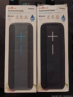 Front view of two Pick-me PK6825 Portable Bluetooth Speakers in boxes, black and gray versions with volume control buttons visible.
