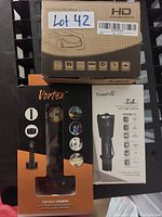 Photo showing one boxed HD car dash camera and two boxed flashlights (Vortex and TrustFire T4) arranged on a black surface.