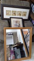 Overall view of two framed art prints and framed mirror leaning on chair and stacked on table.