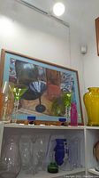 Top shelf with framed colored glass art, green stemmed glass, yellow vase, and a pink item with other glasses below.