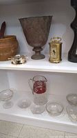 Shelf featuring cranberry glass pitcher grouped with crystal glass candy trays and bowls, large metal vase, small brass clock, woven pottery basket