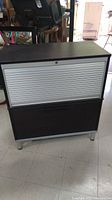 Front view of modern filing cabinet with black wood top and bottom and a silver roller shutter door.