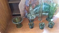 Photo of seven pieces of green glass decor displayed on a wooden surface, showing varying shapes and sizes including pedestal vase, slender vase, and candle holders.