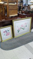 Both floral prints in gold frames leaning against furniture showing size difference and condition.