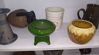 Nine vintage pottery pieces displayed on a shelf including crocks, jugs, and planters.