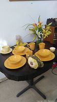 Full set arranged on black table showing yellow dishes, mugs, bowls, teapot, artificial flowers, and decor pieces.