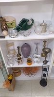 Shelf view showing various decorative trinkets including green ceramic piece, metal sugar bowl, glass candlesticks, brass candlesticks, crystal decanter, small glass dish, silver pitcher, wooden pedestal dish, and woven baskets with dried floral stems.