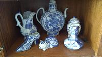 Four white and blue porcelain decorative items arranged on a wooden shelf: pitcher, flask vase, decorative shoe, and cat figurine.