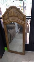 Full view of the rectangular wood framed mirror showing the decorative ornate top and reflection of a chair and door.