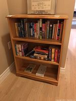 Wooden bookshelf with three shelves filled with books on light wood floor and beige wall