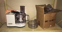 Photo showing Braun food processor main unit with clear plastic mixing bowl and accessories next to a cardboard box containing additional parts and some cookbooks behind it.