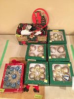 Photo showing five green boxes containing mostly clear and gold Christmas baubles, a small basket with other ornaments inside, and a red and beige Christmas-themed mat underneath.