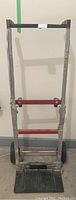 Front view of a metal dolly hand truck with a red and gray frame, standing upright against a wall.