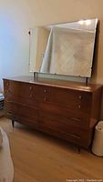 Wooden dresser with attached large beveled mirror, showing six drawers with two different handle styles, standing on four tapered legs.