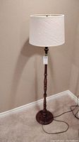 Full view of the floor lamp showing wood stem, round base, and white shade against beige wall.