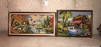 Two framed needle point landscape artworks leaning against a basement wall, one smaller watermill scene on the right, larger dogs and pond scene on left.