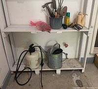 Wide view showing white shelving unit with multiple garden tools including Home Hardware sprayer, galvanized watering can, and hand tools inside green-striped container