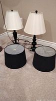 Two black wooden table lamps with distinct bases (square and rounded) with two matching black cylindrical lampshades placed on carpet floor.