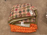 Folded quilt with multicolored circular floral patterns on cream background alongside folded bright orange fringed bedspread.