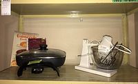 Shelf with Black & Decker electric skillet with glass lid and cord, Sunbeam MixMaster stand mixer with glass bowl and beaters, plus two cookbooks.