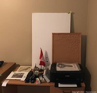 Photo of Canon printer on a wooden desk with documents, manual, and cork board behind it, plus foam display boards and other office supplies around.