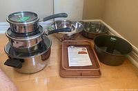 Two sets of stacked stainless steel pots with lids, together with a colander, Bundt pan, angel food pan, crisper tray, and cutting board arranged on floor in corner.