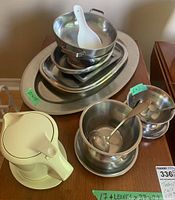 Overview of the complete serving ware set including stacked stainless steel bowls and trays and a white thermal gravy server with a ceramic spoon on top.