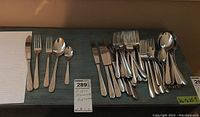 Two sets of stainless steel flatware pieces arranged on a wooden shelf, showing two different handle patterns; includes knives, forks, spoons, and pie forks.