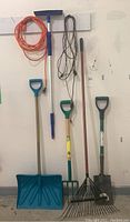 Full view of the seven items hanging on a wall: blue snow shovel, pitchfork, rake, squeegee, and three electric extension cords (orange, brown, and another brown).