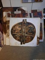 Four unframed paintings stacked leaning against a wall, showing abstract circular motif and human figure paintings.