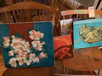 Three 12x10 floral paintings on board displayed on chairs with two other paintings visible nearby.