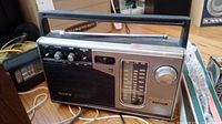 Front and side view of silver Sony TR20 3-band radio showing speaker grille, tuning dial, control knobs, and handle.