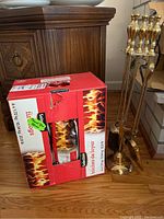 Red and white box showing 6 fire logs, unopened but with torn top flap, next to brass fireplace tools on wooden floor.