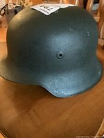Green metal military helmet shown at a slight angle to display the side vent hole and curved shape.
