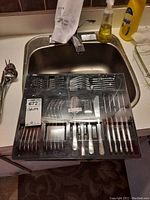 Top view of a black tray containing neatly arranged stainless steel forks, spoons, knives, and cutters at a kitchen sink.