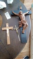 Three crucifixes displayed on a wooden table; one large wood crucifix with dark wood carved figure, one medium wood crucifix with white plastic figure, and one small gold-colored metal crucifix.
