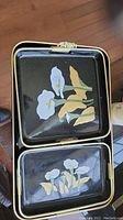 Two square black decorative platters with floral artwork placed one on top of the other
