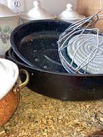 Photo showing black speckled enamel roasting pans stacked with various metal and silicone trivets and a whisk rack on top, white ceramic canisters with lids in background.