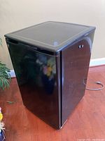 Black compact GE dorm refrigerator shown from an angled side view on hardwood floor with some reflections visible on the surface.