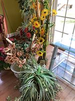 Overall view of multiple faux plants including sunflower and leafy plants with pots and stands near a sliding door