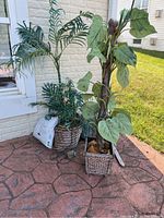 Two faux plants side by side beside a brick wall, showing full plants and pots on a paved surface.