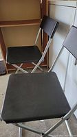 Two matching folding chairs with square seats and backs, metal silver frames, black seats and backs.