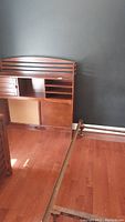 Photo of wooden headboard with storage shelves and part of metal bed frame on hardwood floor against dark wall.