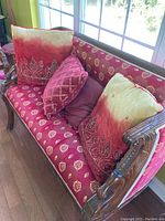 Loveseat with red and gold patterned upholstery, carved wood frame, and multiple decorative pillows visible in natural light near window.