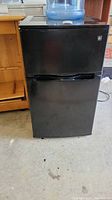 Closed black mini refrigerator/freezer combo with a water jug on top.