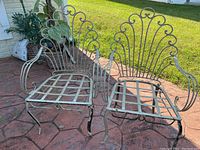 Two white metal outdoor chair frames on a stamped concrete patio, showing decorative scroll designs on the back and arm rests, no cushions present.
